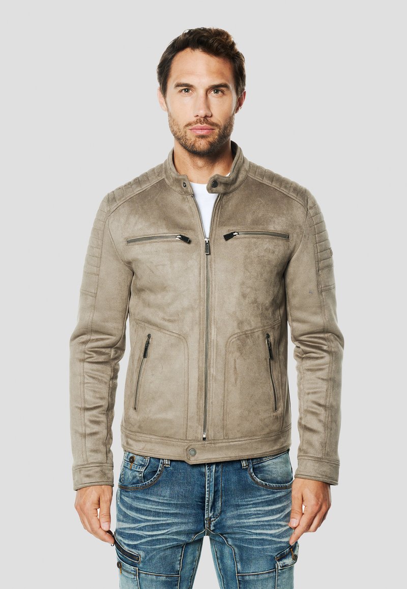 Béžová semišová bunda so stojacím golierom, zipsom na prednej strane a vreckami, rebrovanými detailmi na ramenách a priliehavým strihom. Kombinovaná s modrými rifľami.