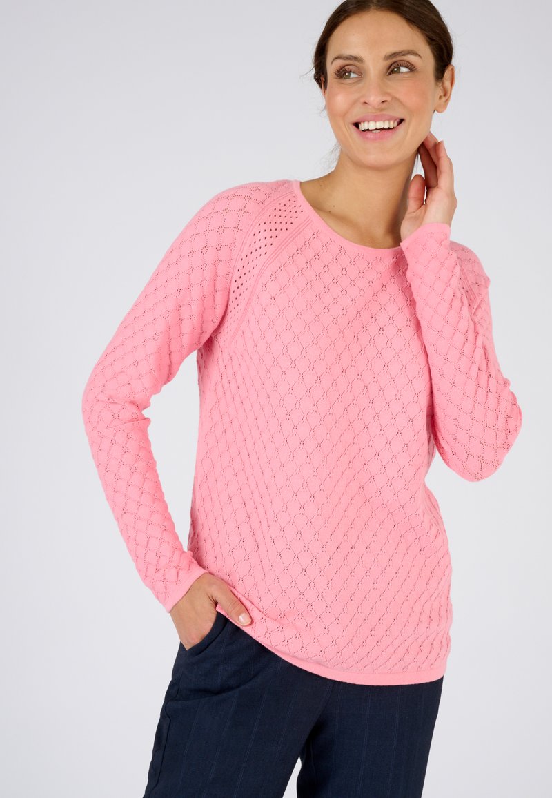 Pull en tricot rose avec un motif en losanges, encolure ronde, manches longues et poches sur les côtés, associé à un pantalon foncé.