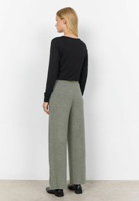 Schwarzes Langarmoberteil kombiniert mit lockeren, hellgrauen Wollmischhosen. Flache schwarze Schuhe runden das Outfit ab. Die Ansicht zeigt die Rückseite des Models.