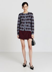 Strukturierter Cardigan mit Rautenmuster in Grau, Burgund und Schwarz; kombiniert mit einem figurbetonten Burgunderrock und schwarzen spitzen Schuhen. Kleine schwarze Tasche.