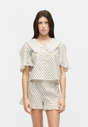 Femme portant un chemisier blanc à manches courtes et un short assorti avec des pois marron foncé et des bords volantés, debout devant un fond uni.