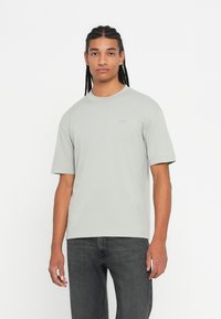 Νεαρός άνδρας με πλεξούδες, φορώντας ελαφρύ γκρι oversized μπλουζάκι και σκούρους γκρι τζιν, όρθιος μπροστά σε απλό λευκό φόντο.