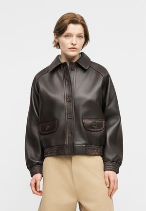 Fiatal személy sötétbarna bőrgombos dzsekit visel, amelyen zsebfedők találhatók, és bézs színű nadrágot, egyszerű háttér előtt áll.