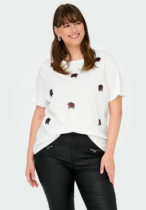 Femme souriante aux cheveux bruns porte un t-shirt blanc avec des imprimés de roses rouges et un pantalon noir avec des poches zippées sur un fond clair.