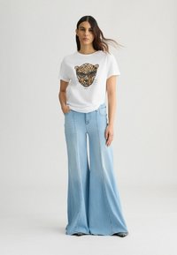 T-shirt bianco con grafica di una faccia di leopardo, abbinato a jeans a gamba larga azzurri. L'outfit ha una vestibilità casual e rilassata.