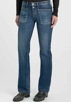 Vrouw draagt blauwe rechte jeans met voorzakken en zwarte loafers, staand tegen een effen lichte achtergrond.