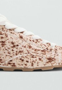 Zapatilla beige con superficie texturizada, que presenta motas marrones, cordones blancos y una suela de goma marrón. Detalles de costura visibles a lo largo de las costuras.