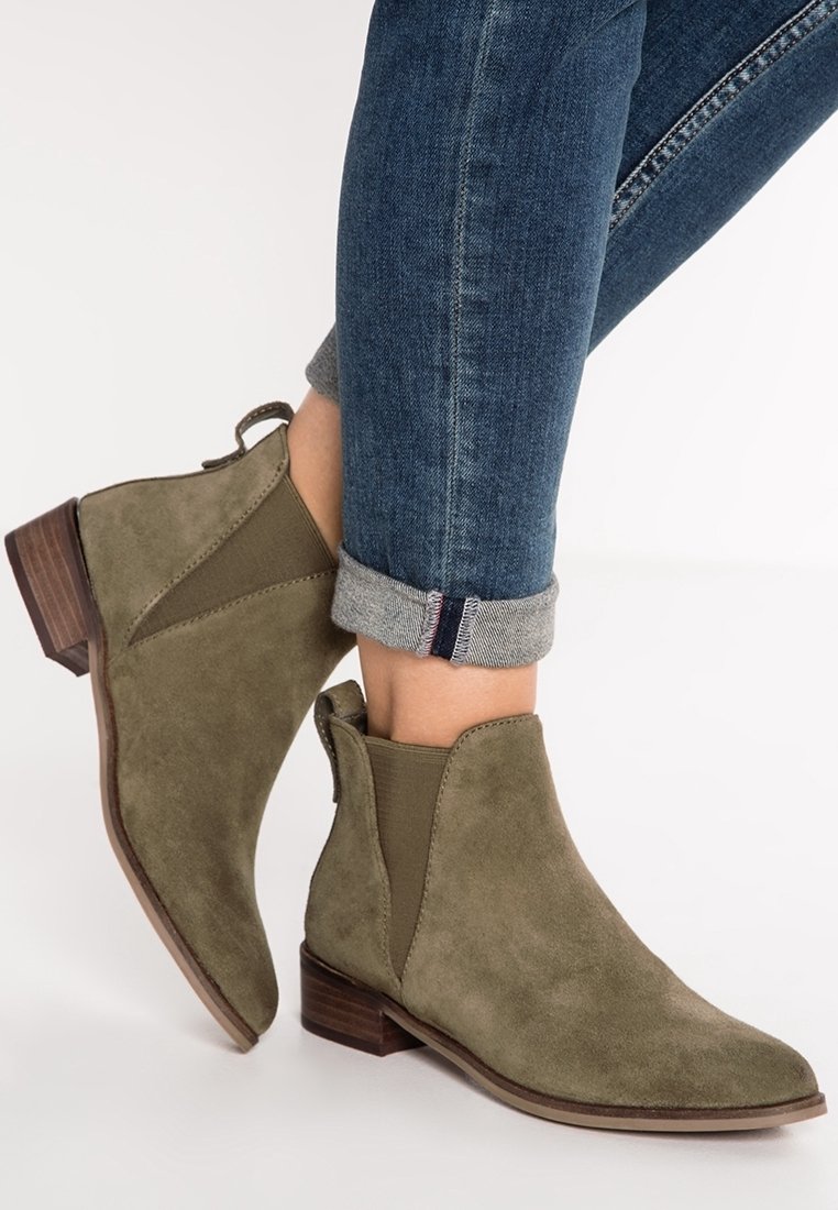 Bottes cheville en daim vert olive avec panneaux latéraux élastiques et un petit talon en bois empilé, portées avec un jean en denim retroussé.