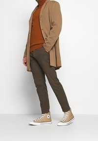 Béžový svetr na zapínání přes hlavu přes hnědý rolák, kombinovaný s olivově zelenými kalhotami a béžovými teniskami s vysokým svrškem. Svetr má pohodlný střih.