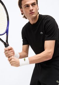 Νεαρός άντρας σε μαύρη αθλητική φόρμα κρατώντας μια ρακέτα του τένις, φοράει λευκά wristbands, προετοιμάζεται να χτυπήσει μια μπάλα του τένις με συγκεντρωμένη έκφραση.
