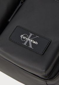 Czarna nylonowa torba o teksturowej powierzchni, z prostokątną naszywką z logo Calvin Klein oraz zamknięciem na zamek. Prosty design bez dodatkowych akcentów.