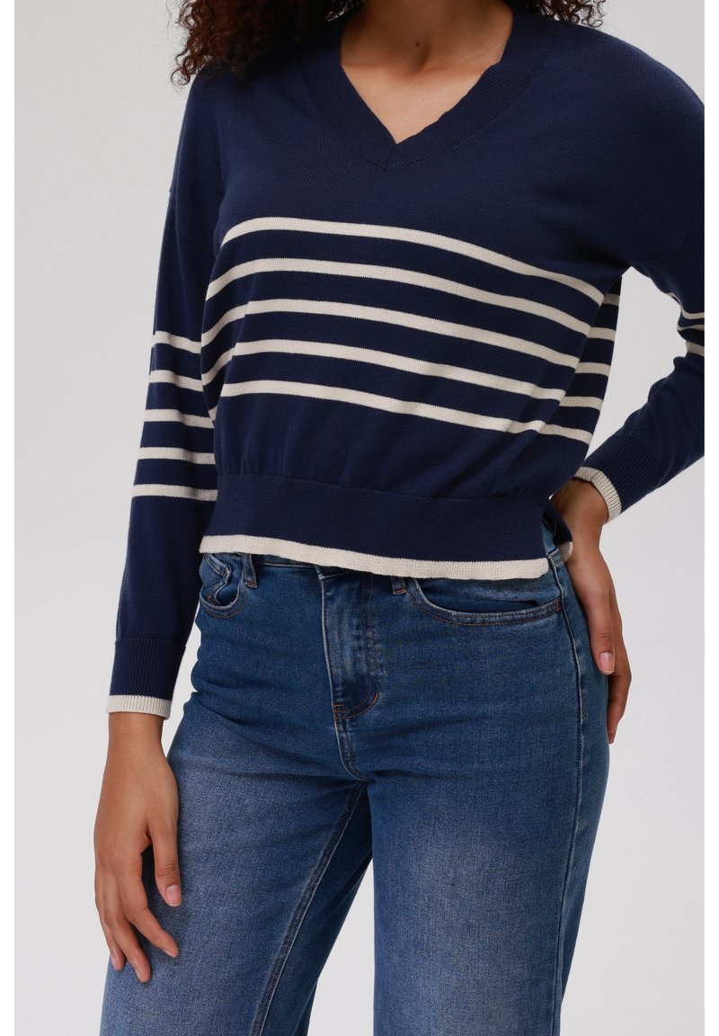Femme portant un pull col V bleu marine avec des rayures horizontales blanches et un jean bleu taille haute délavé moyen, main posée sur la hanche.