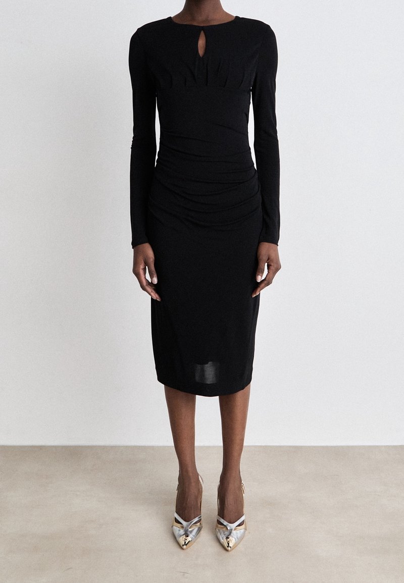 Femme portant une robe midi noire cintrée à manches longues avec un détail œillet sur le devant, associée à des escarpins métalliques à bout pointu.