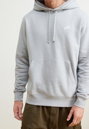 Personne portant un sweat à capuche Nike gris clair avec cordons et poche avant, associé à un pantalon marron foncé, sur un fond uni.