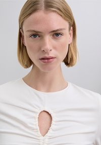 Top de algodón blanco con diseño ajustado, que presenta una sección central fruncida y un corte en forma de ojo de cerradura en el pecho. Textura suave.