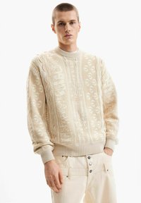 Fiatal férfi rövid hajjal, bézs sz ínű, mintás kötött pulóvert és világos színű nadrágot visel, egyik kezét a zsebében tartva áll, fehér háttér előtt.