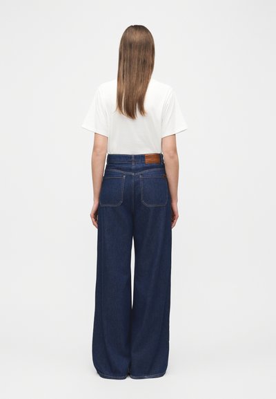 Άτομο με μακριά καστανά μαλλιά, φορώντας λευκό μπλουζάκι και σκουρόχρωμο μπλε τζιν παντελόνι φαρδύ στο κάτω μέρος με ψηλή μέση, στέκεται με την πλάτη γυρισμένη.