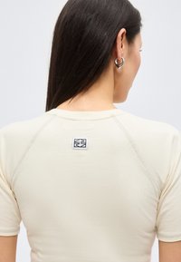 Γυναίκα με μακριά σκουρόχρωμα μαλλιά, που φοράει ένα κρεμ κοντομάνικο πουκάμισο με μικρό ασπρόμαυρο λογότυπο στο πάνω μέρος της πλάτης και ασημένια σκουλαρίκια κρίκους.