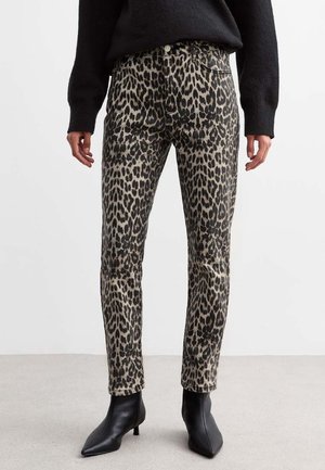 Femme portant un pantalon slim taille haute à imprimé léopard, un pull noir et des bottines noires à bout pointu, debout sur un sol gris.