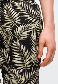 Gros plan de la main d'une personne reposant à côté d'un pantalon noir avec un motif beige de feuilles de palmier tropical.