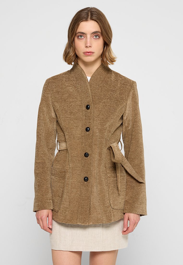 Giacca marrone con texture, colletto alto, design a un bottone, tasche frontali e dettaglio in vita legato. Bottoni neri contrastano con il tessuto.