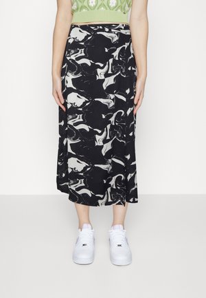 Personne portant une jupe midi à motif abstrait noir et blanc, un crop top vert et des baskets blanches, debout devant un fond blanc uni.