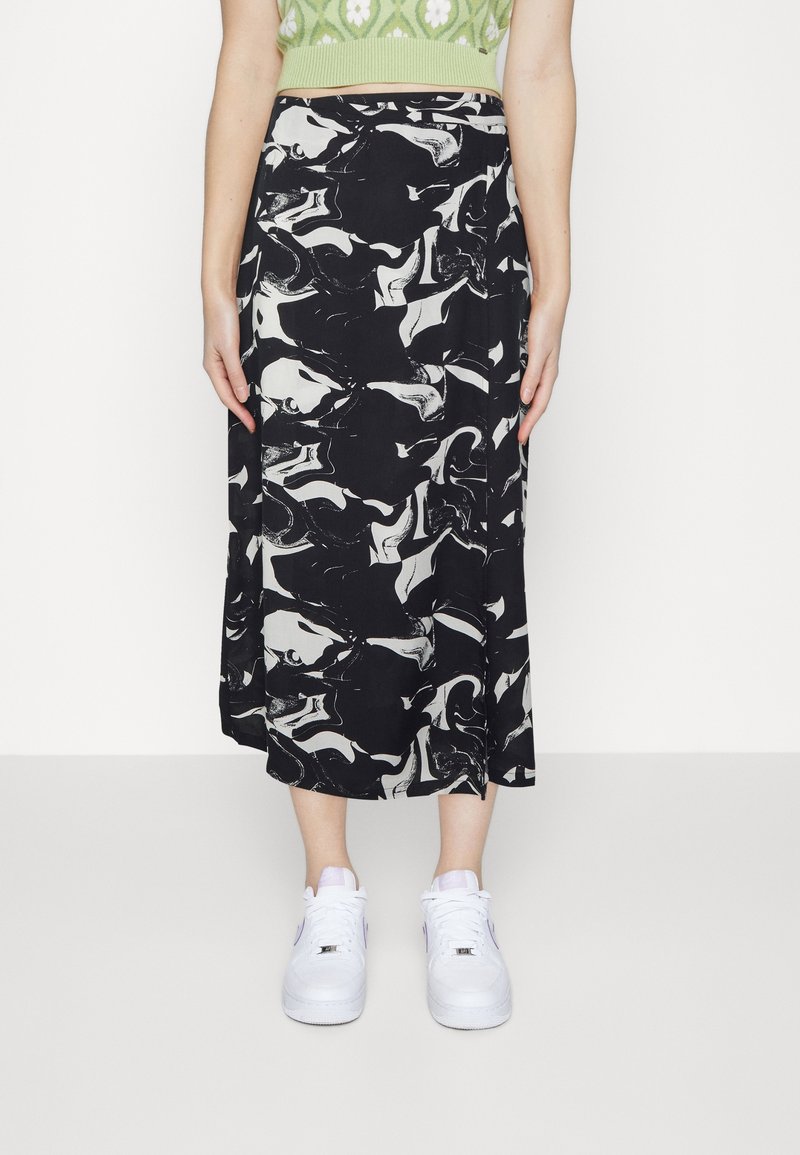 Personne portant une jupe midi à motif abstrait noir et blanc, un crop top vert et des baskets blanches, debout devant un fond blanc uni.