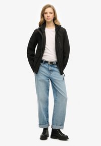 Sort vandtæt jakke med hætte, båret over en hvid t-shirt. Løse lyseblå jeans med optrukne kanter og sorte snørebåndsstøvler.