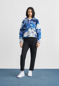 Ung kvinde står på blåt tæppe iført en blå og hvid mønstret fleece, sorte bukser og hvide sneakers mod en ensfarvet hvid væg.