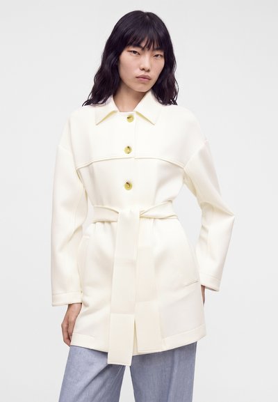 Femme aux cheveux noirs portant un manteau ceinturé crème avec de gros boutons et un pantalon bleu clair sur un fond uni.