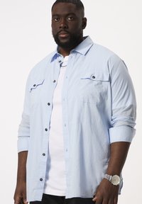 Man met een lichtblauw overhemd over een wit t-shirt, donkere broek, zilveren horloge en kettinghalsketting, staand tegen een witte achtergrond.