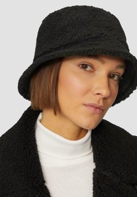 Chapeau seau noir en matériau polaire texturé, avec un large bord et une apparence douce et confortable. Porté avec un col roulé blanc.