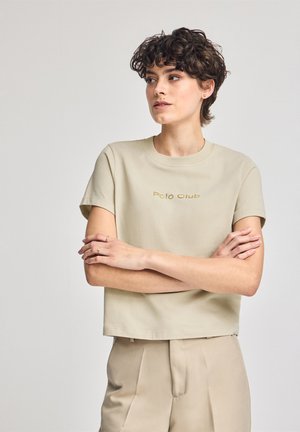 Beige cropped t-shirt med korte ærmer og rund halsudskæring, med et guld "Polo Club" print over brystet. Glat bomuldsstruktur.