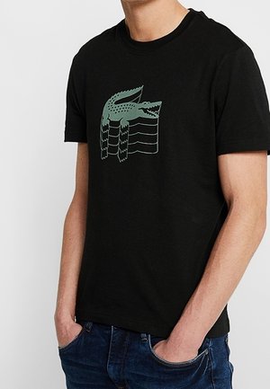 T-shirt noir avec un motif stylisé de crocodile vert sur la poitrine, porté par une personne les mains dans les poches d'un jean bleu.