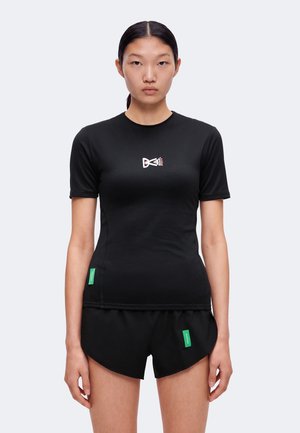 Femme aux cheveux noirs et raides porte un T-shirt noir moulant et un short noir avec des étiquettes vertes, debout devant un fond clair uni.