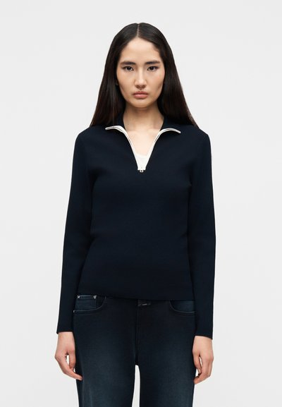 Femme portant un pull noir à col zippé par-dessus un haut blanc et un jean bleu foncé, debout devant un fond clair uni.