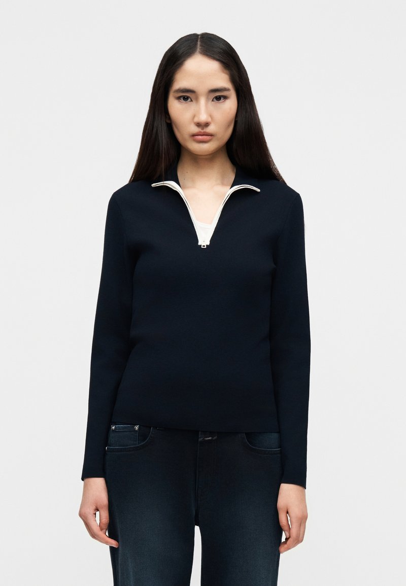 Γυναίκα που φοράει μαύρο πουλόβερ με φερμουάρ στον λαιμό πάνω από λευκό τοπ και σκουρόχρωμο μπλε τζιν, στέκεται μπροστά σε απλό ανοιχτό φόντο.