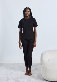 Fekete rövid ujjú pamut póló piros "HUGO" logóval, fekete slim fit nadrággal párosítva. A modell mezítláb áll egy világos szőnyegen.