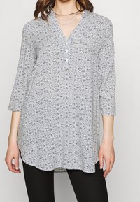 Kobieta ubrana w długą, wzorzystą tunikę z guzikami i dekoltem w serek, połączoną z czarnymi spodniami i warstwowymi srebrnymi naszyjnikami.