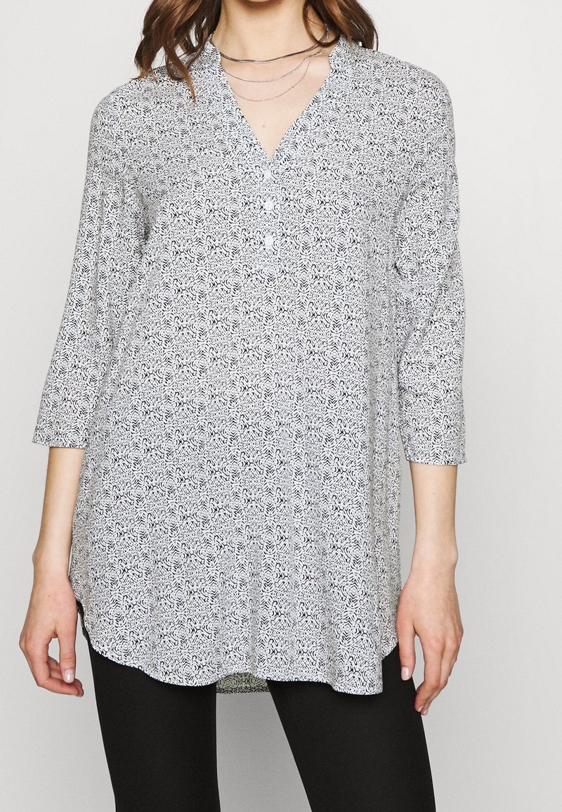 Kobieta ubrana w długą, wzorzystą tunikę z guzikami i dekoltem w serek, połączoną z czarnymi spodniami i warstwowymi srebrnymi naszyjnikami.