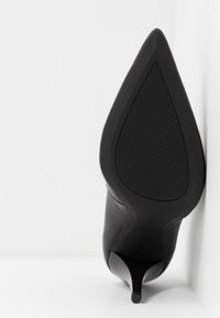 Chaussure à talon haut noire à bout pointu, posée sur le talon contre un mur d'angle blanc, avec la semelle texturée visible.