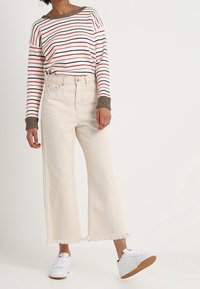 Haut à manches longues rayé avec des lignes rouges et bleu marine, jean beige à jambes larges avec ourlet effiloché, et baskets blanches avec une semelle marron.