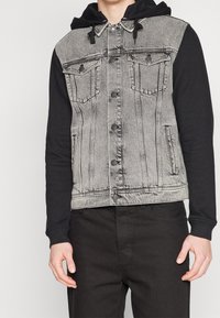 Man i en grå denimväst med svarta knappdetaljer över en svart huva och svarta byxor, som står mot en enfärgad bakgrund.