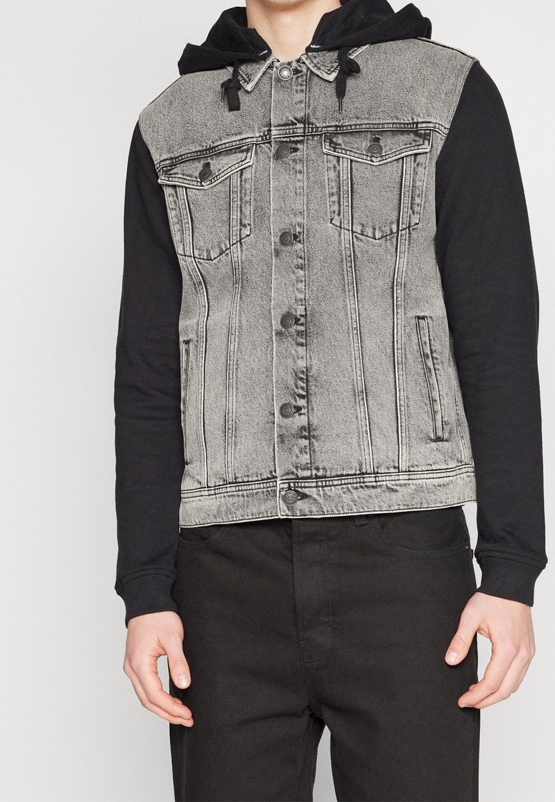 Man i en grå denimväst med svarta knappdetaljer över en svart huva och svarta byxor, som står mot en enfärgad bakgrund.