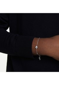 Bracelet en argent avec un charme circulaire et deux accents cylindriques suspendus, se détachant sur un pull sombre et texturé.
