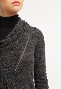 Šedý pletený svetr s volným límcem, který má stříbrný zip na boku a měkký, strukturovaný povrch.