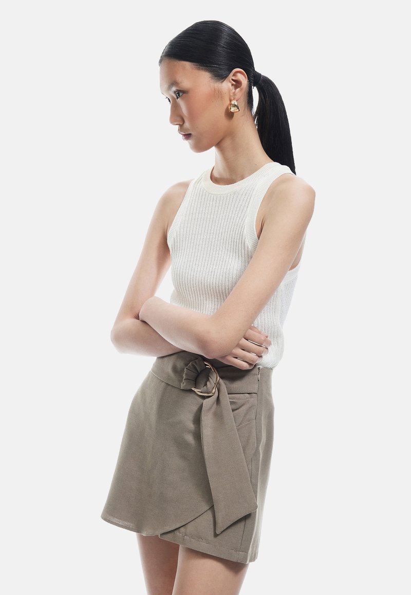 Top sin mangas de color blanco con textura acanalada, combinado con una falda envolvente de color caqui que presenta un lazo atado y un detalle en metal circular. Textura suave, diseño ajustado.