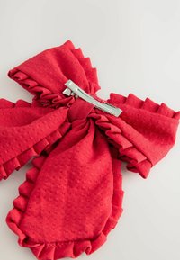 Un nœud en tissu rouge avec des bords froncés, fixé par une pince en métal, repose sur un fond clair, mettant en valeur sa texture et sa couleur vibrante.