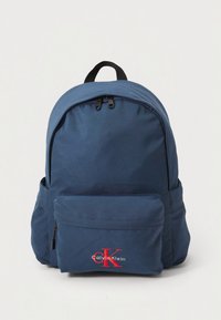 Navy blue nylon backpack with a rounded design, dual zippers, front pocket, and a red Calvin Klein logo on the lower front.