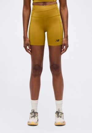 Femme portant un short cycliste taille haute jaune moutarde et des chaussures de sport blanches avec des chaussettes, debout devant un fond uni.